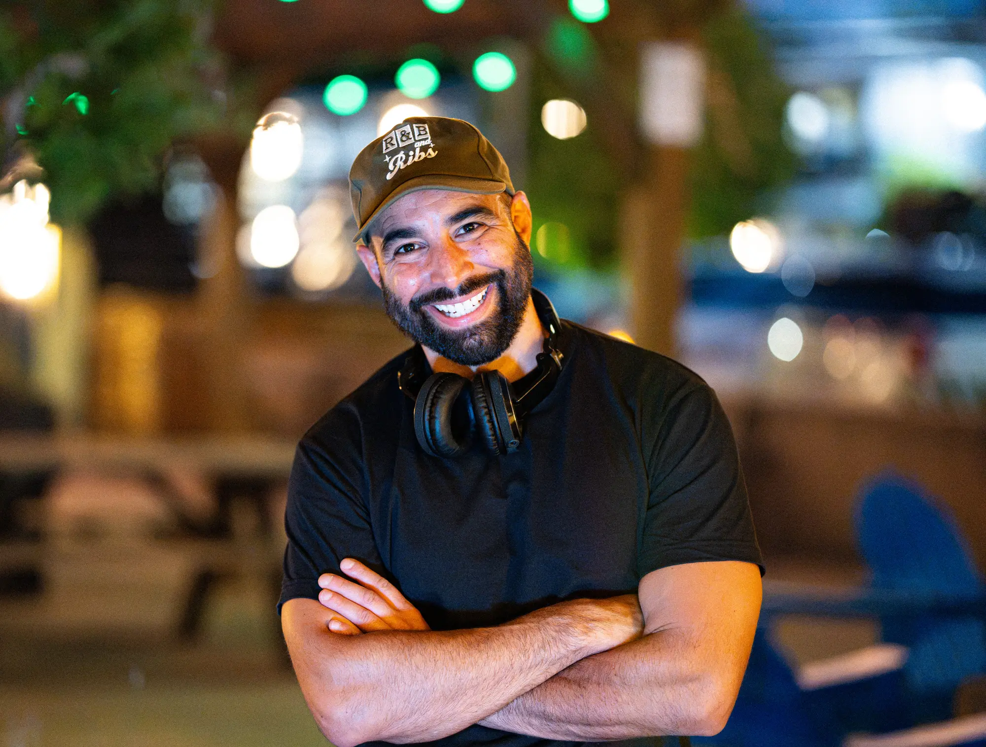 Portrait of DJ Mojo in a cap and black t-shirt, smiling with his arms crossed and headphones around his neck, in an evening patio with string lights.