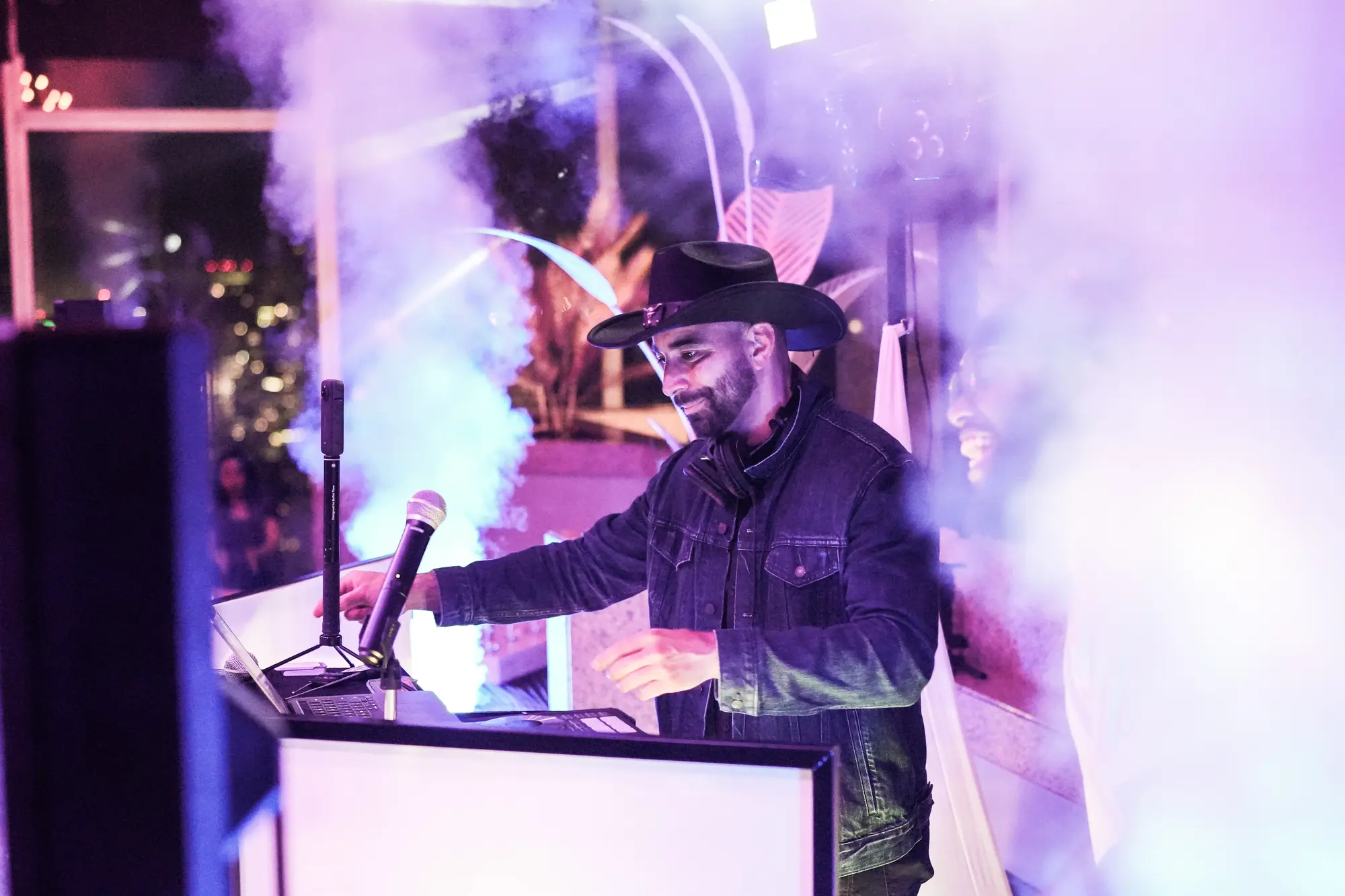 DJ Mojo in a black cowboy hat and denim jacket, smiling at the decks with a microphone, in a cloud of purple haze.