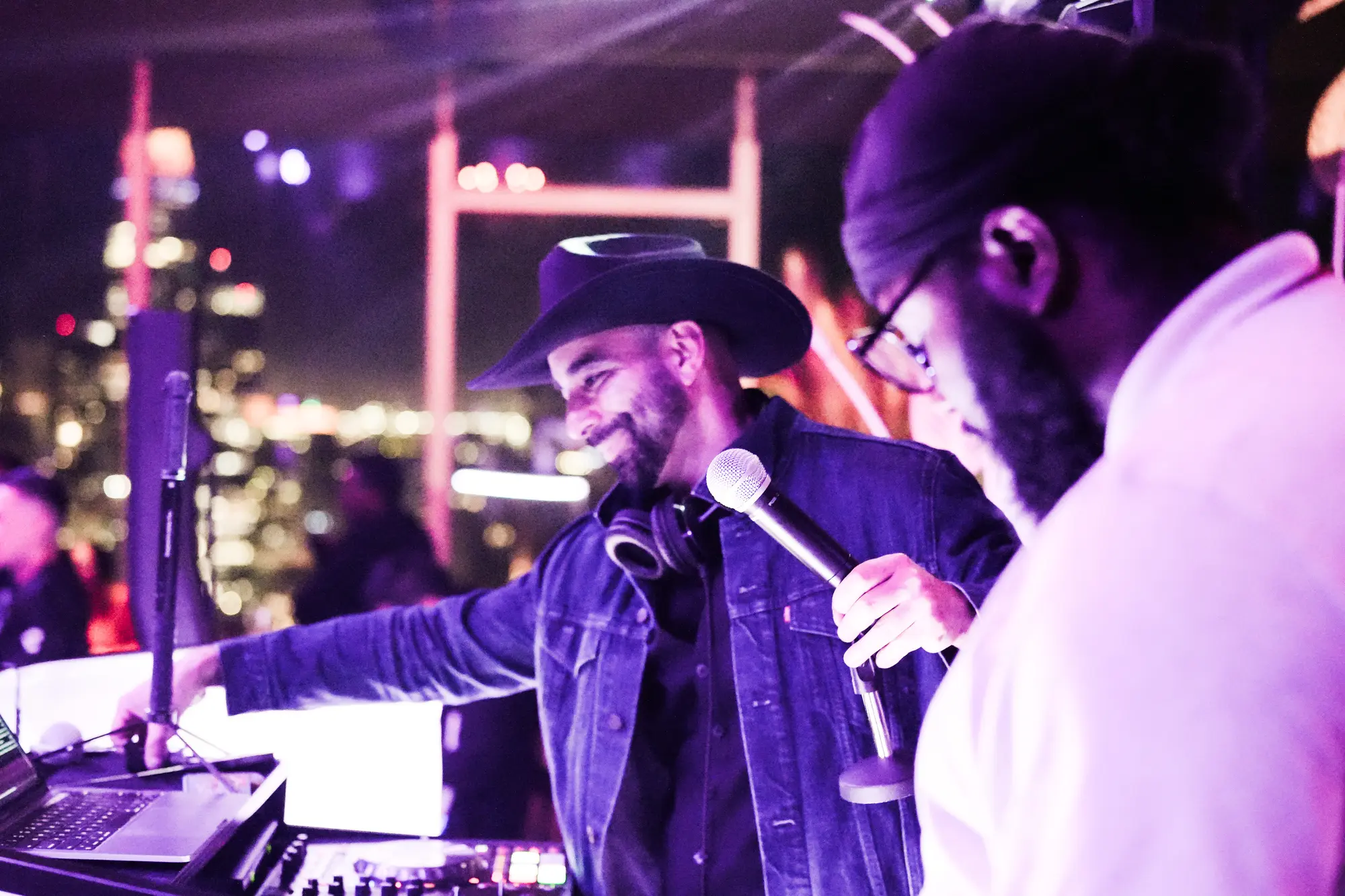 DJ Mojo in a cowboy hat and denim jacket, smiling, with a microphone in hand as he works the mixer under purple light.