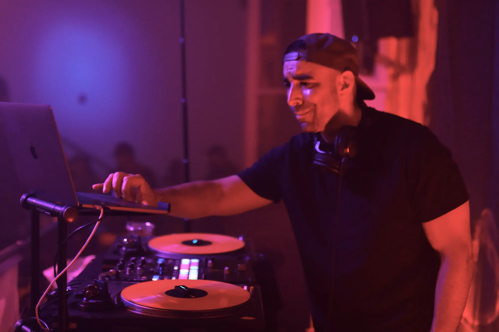 DJ Mojo smiling at his turntables and laptop in a pink and violet lit lounge.