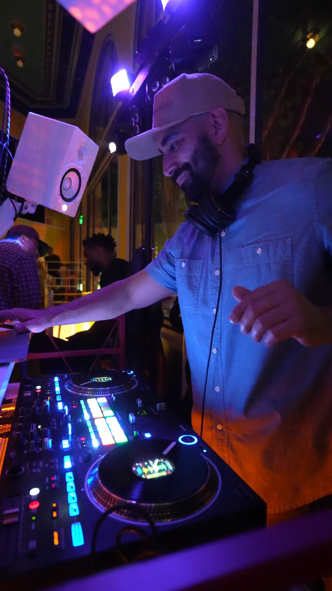 DJ Mojo in a cap and light blue shirt smiling as he works the controller, with colored stage lights around the booth.