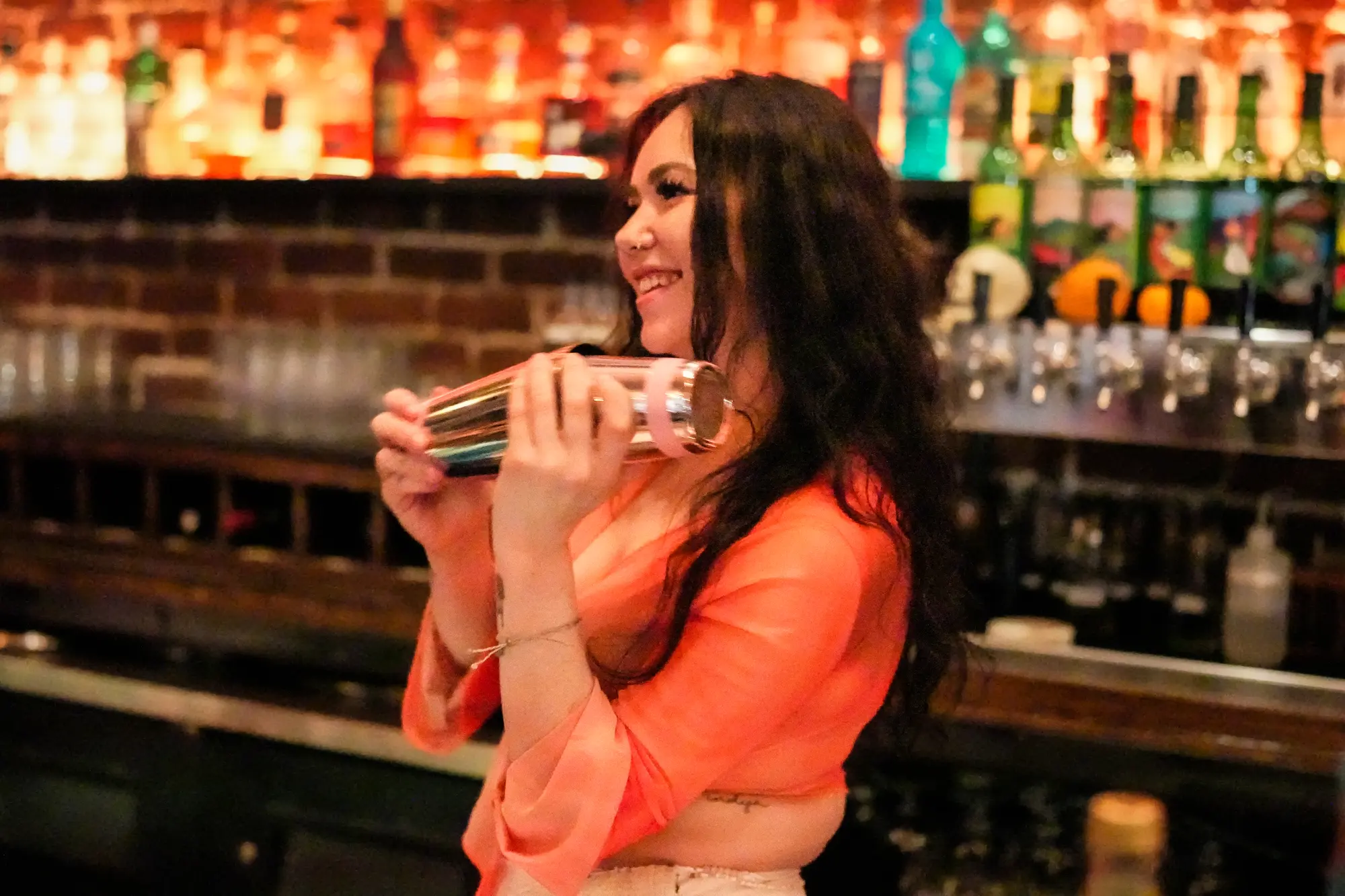 A smiling bartender shakes a cocktail in front of a back bar lit in warm orange.