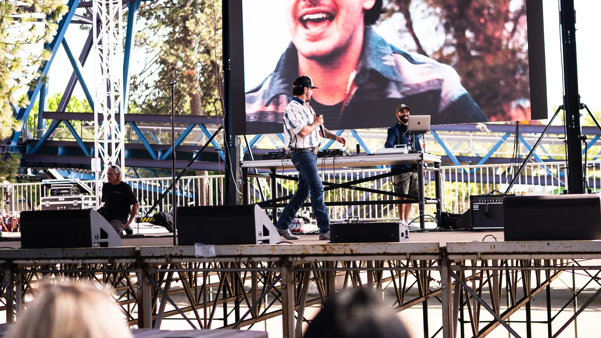 An artist in a cap and striped shirt performs with a microphone on an outdoor stage while DJ Mojo plays from a laptop at a table behind him, a big screen above them.