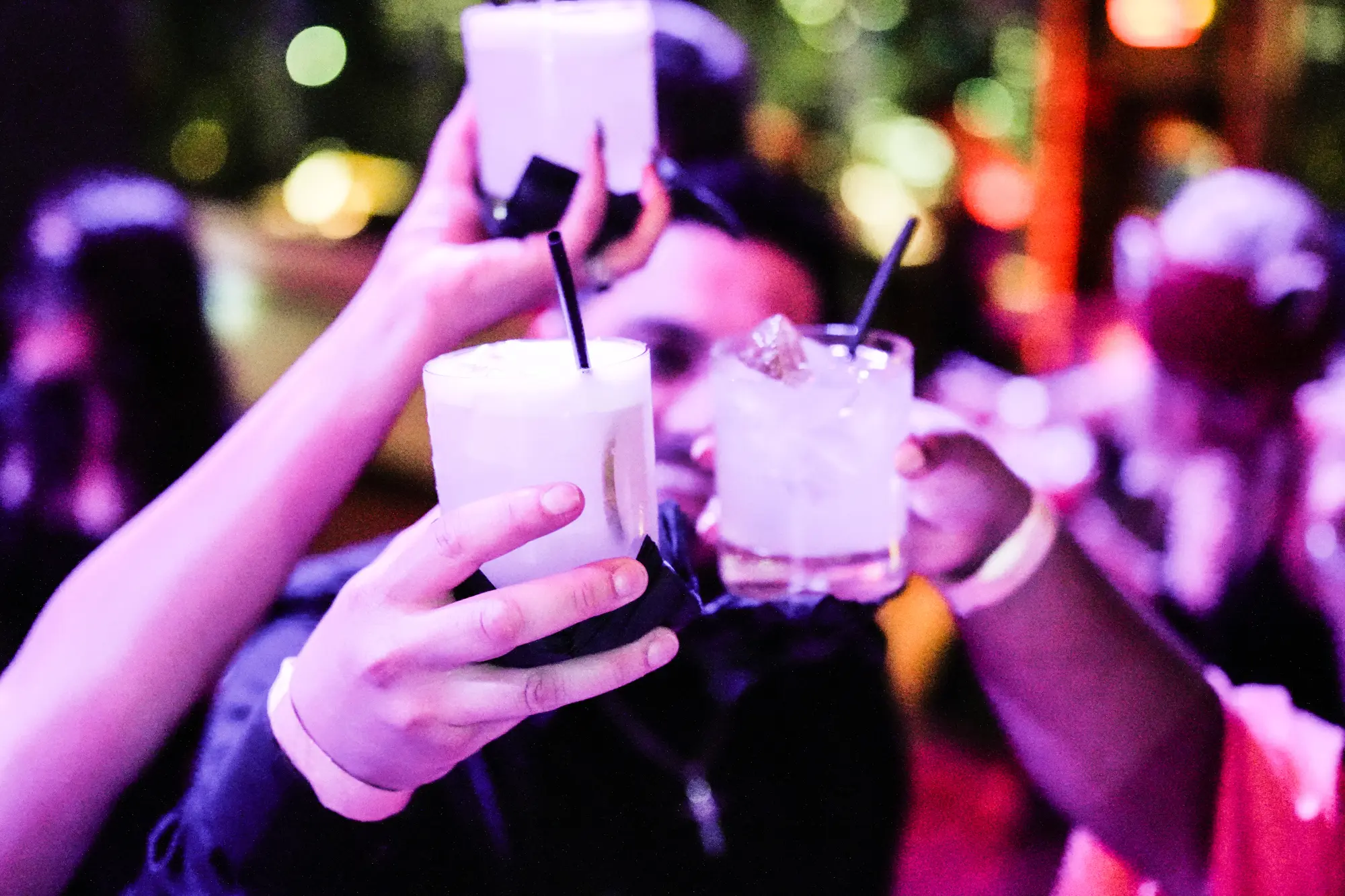 Guests raise three cocktails together in a toast, lit in purple and pink at a party.