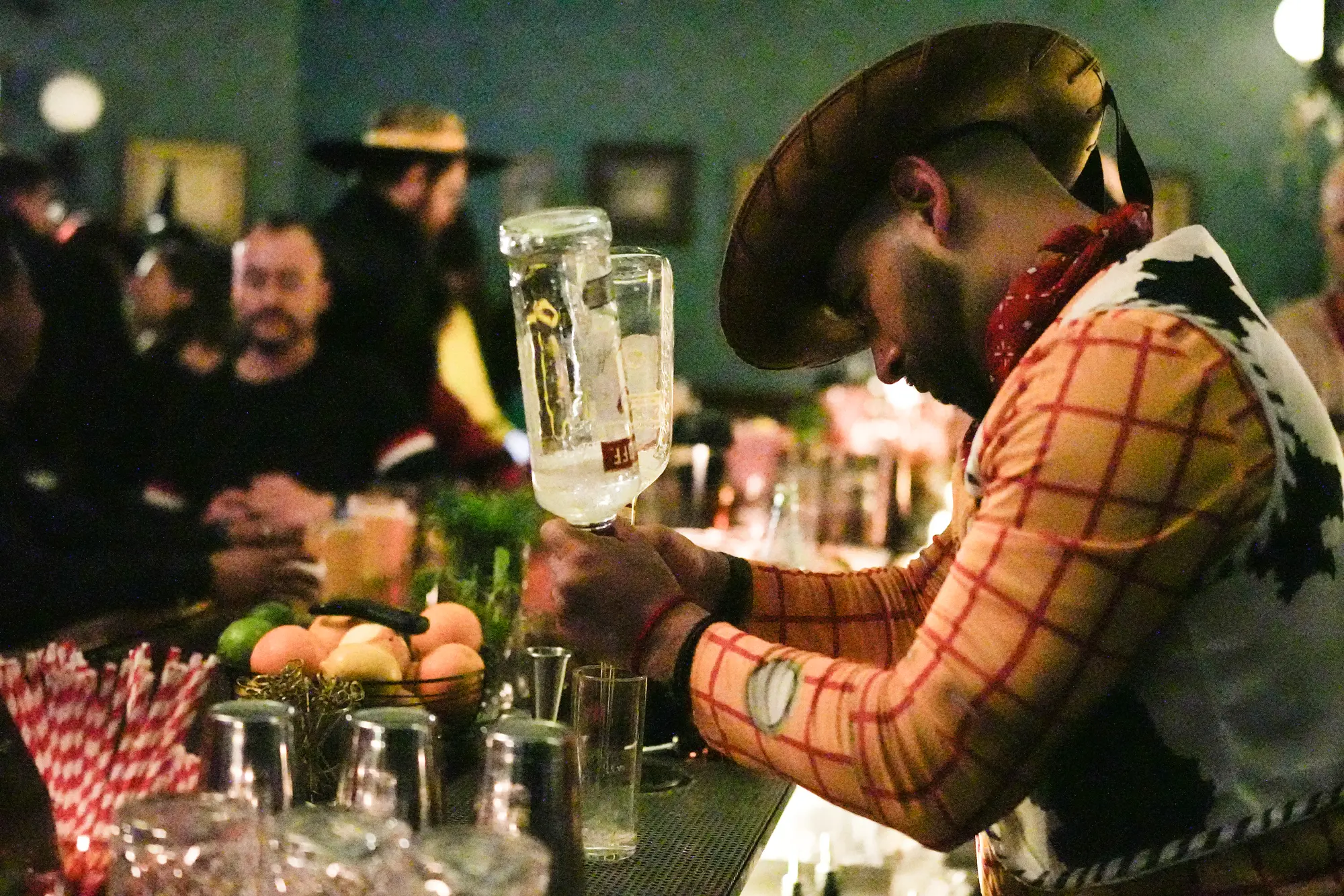 A bartender dressed as a cowboy leans over the bar and pours from a bottle into glasses.