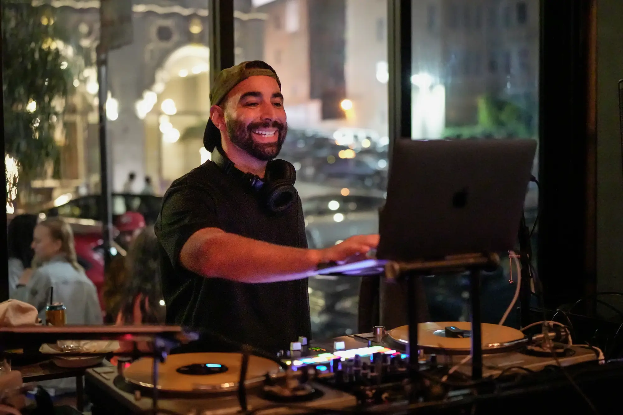 DJ Mojo smiling behind his laptop and turntables at a San Francisco bar, with a lit street window behind him.