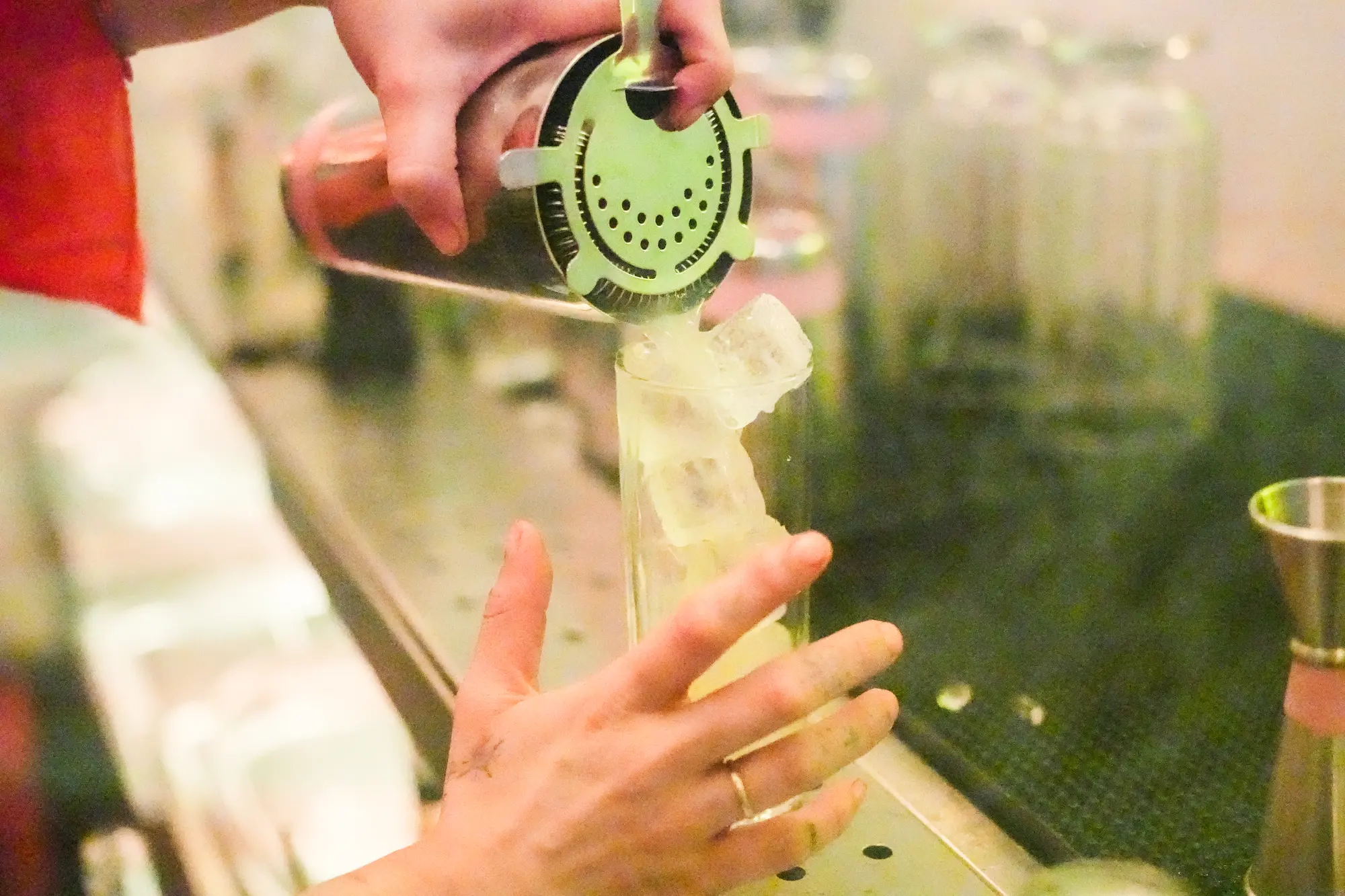 Hands strain a cocktail from a metal shaker through a green strainer into a glass on the bar.