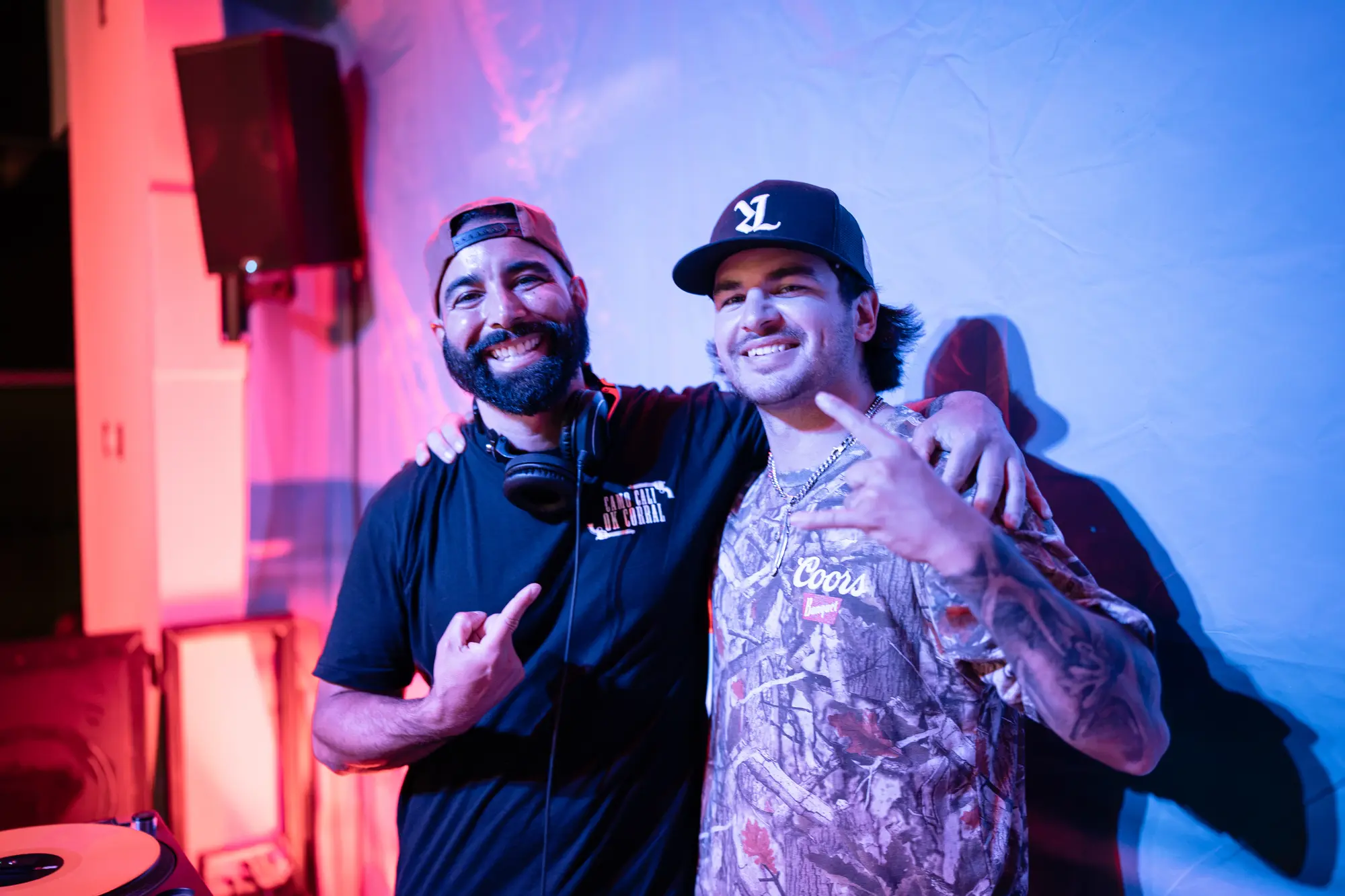 DJ Mojo, headphones around his neck, arm in arm with a guest artist in a cap and camo shirt